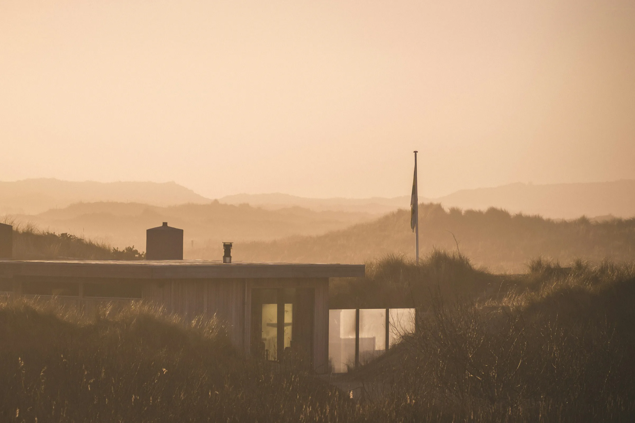 A long timber house at dusk, its windows lit, with the dunes behind
